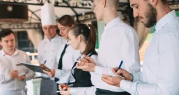 A group of restaurant servers and a chef standing in a line during a training session, taking notes on notepads and clipboards.