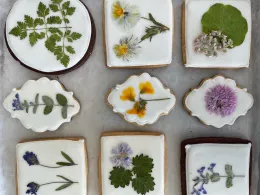 Nine cookies arranged in a grid, each decorated with pressed flowers and leaves on white icing. The cookies vary in shape and feature floral designs.