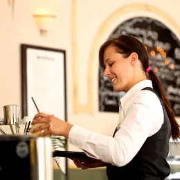 waitress with long brown hair makes coffee wearing black vest and white long sleeve shirt
