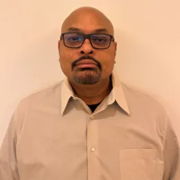 Derrick Tiwari, Assistant Director of Admissions, looks at the camera in front of a tan wall.