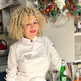 Georgianna Hiliadaki is an ICE alumni. She is standing in a white chef's coat in a kitchen area.
