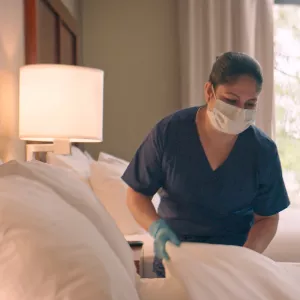 A housekeeper makes a hotel bed with a face mask on