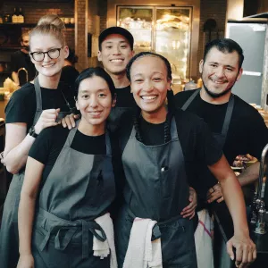 A group of ICE Los Angeles alumni in black shirts and blue aprons at the Otium restaurant kitchen smile