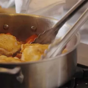Tongs grab a piece of chicken being cooked as coq au vin in a steel pan