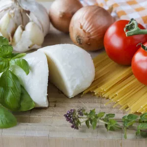 Fresh mozzarella, tomatoes, onions, pasta and garlic sit on a wooden surface