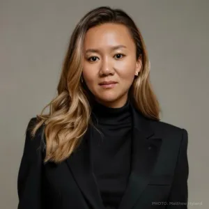 Simone Tong is an ICE alum. She is standing in a black blazer in front of a tan wall. 