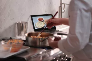 A student cooks a dish for ICE's online Culinary Arts program while watching an instructional video on a laptop