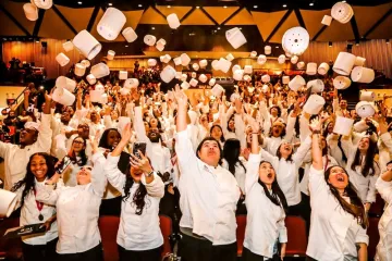 A large group of graduating culinary students wearing white chef coats celebrate inside an auditorium. They are cheering, smiling, and throwing their tall white chef hats (toques) high into the air.