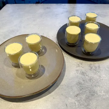 Eight glasses of yellow lassi sit on two serving trays on a marble table. 
