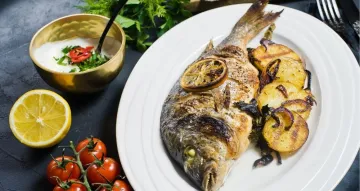 A whole grilled fish served on a white plate with charred lemon slices, grilled potatoes, and red onions, alongside a side sauce, a fresh lemon half, and cherry tomatoes.