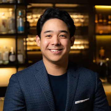 Jhonel Faelnar, Bev Director at NAE:UN Hospitality, smiles wearing navy blue blazer and black t-shirt standing in front of golden lit shelves full of beverages.  