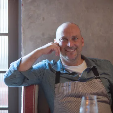 Chef Steve Samson, owner of LA's Rossoblu restaurant, sits in booth with his arm propped on the booth's ledge wearing blue shirt and gray apron.