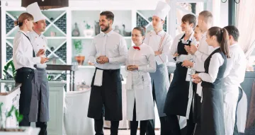 A group of restaurant staff, including chefs in white hats and servers in aprons, standing together and talking in a bright dining room.