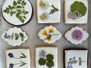 Nine cookies arranged in a grid, each decorated with pressed flowers and leaves on white icing. The cookies vary in shape and feature floral designs.