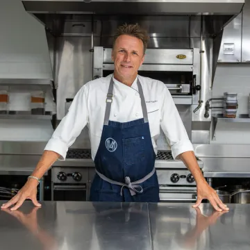 Chef Marc Murphy in chef whites and navy apron stands at counter in industrial kitchen.