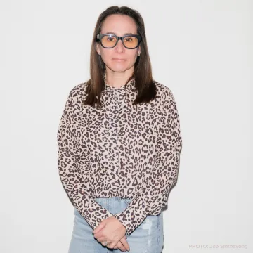 Rebecca Roth Gullo in an animal print top and ripped blue jeans in front of a beige wall.