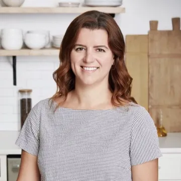 ICE Alumni Sarah Entwistle smiles at the camera in a striped shirt, standing in a kitchen with light-wood cabinets and white plates on shelves