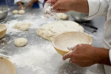 A baker sprinkles flour over the prep table before shaping bread dough