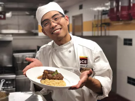 An ICE culinary school in Los Angeles student smiles while holding a plate of bone marrow risotto and meat