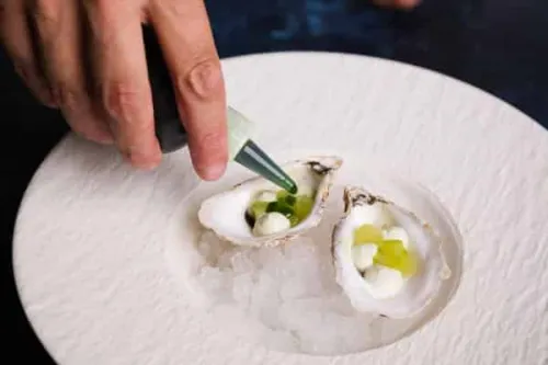 a hand uses a small squeeze bottle of green herb oil onto the bed of an oyster
