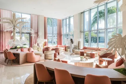 a blurry woman in a white suite walks through a large room with large windows as walls filled with pink furniture and white faux palm trees