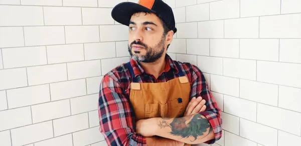 Chef David Viana stands in baseball cap, checkered shirt and leather apron in front of white tiled wall.