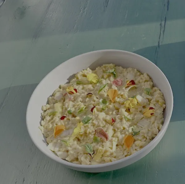 A creamy, light-colored risotto served in a white ceramic bowl, garnished with delicate, colorful edible flower petals and small green herbs. The bowl sits on a rustic, light-teal wooden surface illuminated by soft, natural sunlight from the upper left.