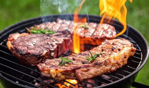 Thick steaks grilling over an open flame on a charcoal barbecue, seasoned with peppercorns and fresh rosemary.