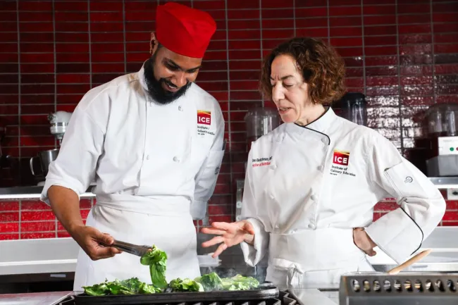A student practices grilling bok choy while his instructor looks on