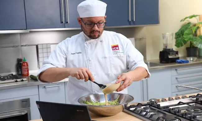 A student in an ICE white uniform pours a beige sauce into a bowl of greens for plant based culinary school online