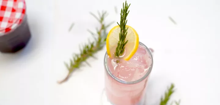 A pink drink garnished with rosemary and lemon on a white table