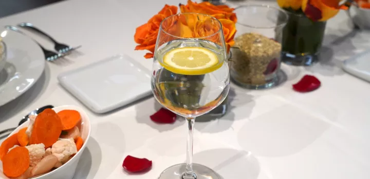 A wine glass filled with water and a slice of lemon sits on a table with a white table cloth and orange flowers