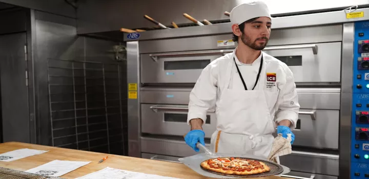 An ICE student holds a pizza on a pizza peel in front of ovens