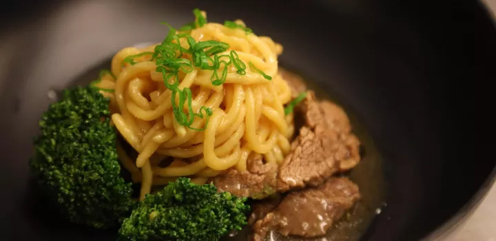 A dish of egg noodles with beef and broccoli sits in a black bowl
