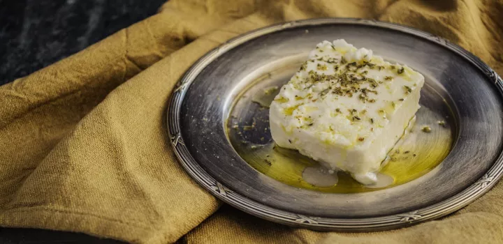 A block of feta cheese with oil and herbs sits on a metal plate on top of a yellow cloth