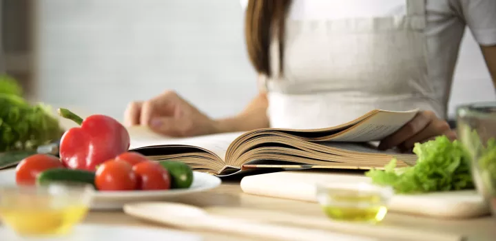 A woman reads a cookbook at a counter with ingredients.