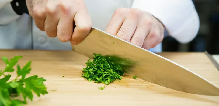 a chef's hands use a knife to cut cilantro on a wooden cutting board