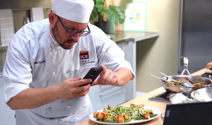 A student in a white ICE coat and hat takes a photo of a plant-based meal for online plant-based culinary school